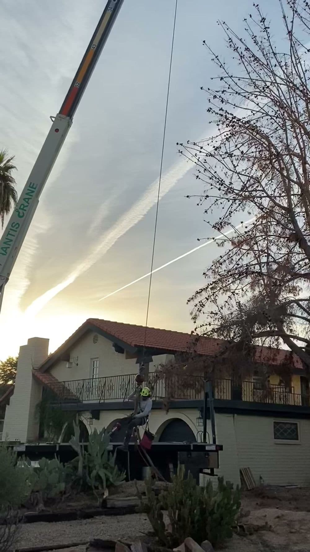 Crane lifting a worker near a house at sunset with jet trails in the sky.