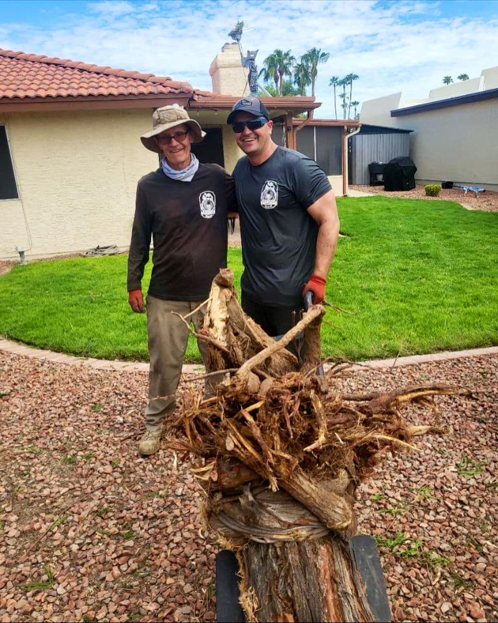 Two men working outdoors, smiling while transporting tree roots on a wheelbarrow.